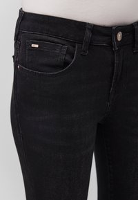Gros plan sur la taille et la poche d'un jean en denim noir, avec passants pour ceinture, bouton métallique et petite étiquette de marque sur la poche avant.