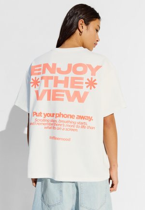 Femme aux longs cheveux foncés porte un t-shirt blanc avec un texte rose « ENJOY THE VIEW » et un message incitant à ranger son téléphone, associé à un jean bleu clair.