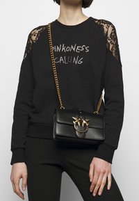 Sudadera negra con detalles de encaje, que presenta "PINKNESS CALLING" en rhinestones. Un pequeño bolso negro con un cierre de ave dorado y cadena.