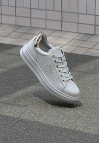 Weiße Ledersneaker mit goldenen Beschlägen, einer strukturierten goldenen Akzentuierung an der Ferse und einem gepolsterten Kragen. Runde Zehenpartie und flache Sohle.