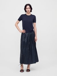 Femme aux cheveux courts et foncés, debout, main sur la hanche, portant un haut bleu marine à manches courtes, une longue jupe plissée avec des sequins, et des talons ouverts noirs.