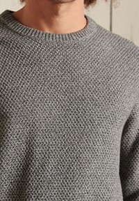 Nahaufnahme einer Person, die einen strukturierten grauen gestrickten Rundhalsausschnitt-Pullover trägt, wobei Oberkörper und Hals gezeigt werden.