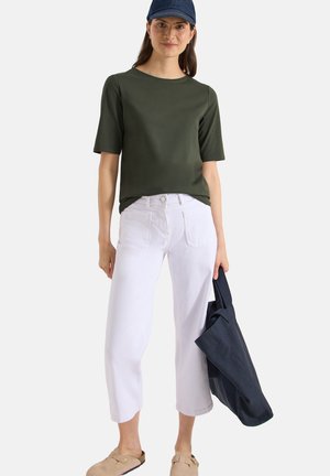 Femme portant un t-shirt vert foncé à manches courtes, un pantalon blanc court, des sandales beiges, une casquette marine, tenant un sac cabas marine sur fond blanc.
