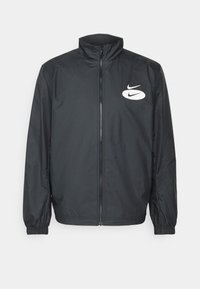 Zwarte Nike windjack met hoge kraag, volledige rits aan de voorkant, elastische manchetten en een wit logo op de linkerborst. Soepele stoftextuur.