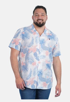 Homme portant une chemise hawaïenne à manches courtes boutonnée, ornée de motifs floraux bleus et roses, associée à un jean bleu, souriant.