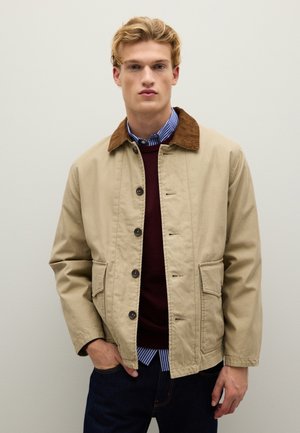Beige Baumwolljacke mit braunem Fischgrätenkragen, vorderen Knöpfen, zwei Seitentaschen, über einem kastanienbraunen Pullover und einem gestreiften Hemd darunter.