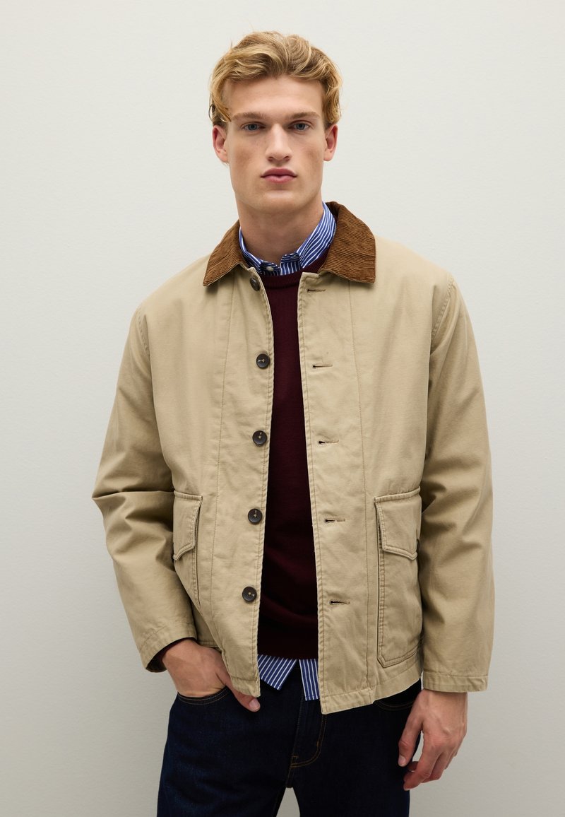 Giacca di cotone beige con colletto in velluto a coste marrone, bottoni frontali, due tasche laterali, indossata sopra un maglione bordeaux e una camicia a righe sotto.