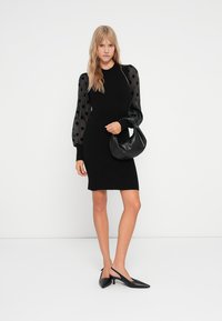 Abito nero con corpetto aderente e maniche a sbuffo in chiffon a pois. Abbinato a una borsa nera e scarpe con punta nera.