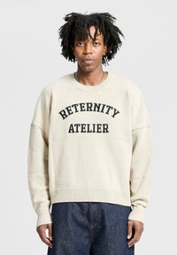 Beige knit sweater with a ribbed hem and cuffs, featuring a black print that reads "RETERNITY ATELIER" across the chest.