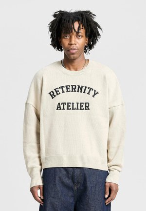Beige knit sweater with a ribbed hem and cuffs, featuring a black print that reads "RETERNITY ATELIER" across the chest.