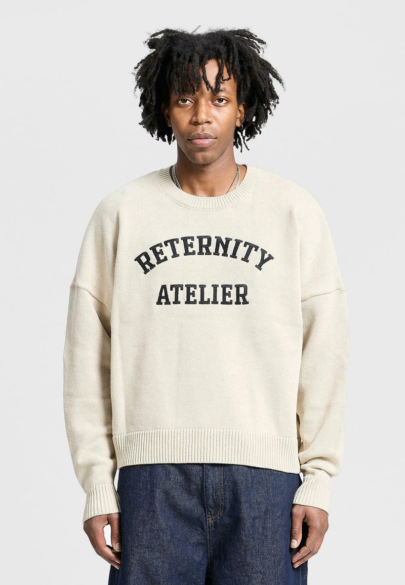 Beige knit sweater with a ribbed hem and cuffs, featuring a black print that reads "RETERNITY ATELIER" across the chest.