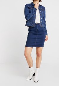 Jeansjacka och kjolset i mörkblått. Jackan har knäppning med knappar och fickor. Vit topp under, tillsammans med vita ankelboots.