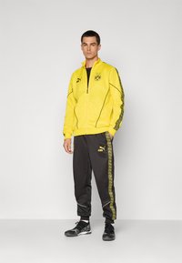 Mand står iført en gul Borussia Dortmund træningsjakke og sorte Puma træningsbukser med gule sidestreger samt sorte sneakers.