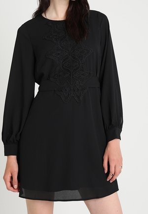 Frau trägt ein langärmeliges schwarzes Kleid mit Spitzenapplikation auf der Vorderseite und steht vor einem einfarbigen hellen Hintergrund.