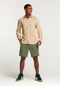 Camicia beige a maniche lunghe con bottoni, pantaloni corti verdi a fantasia con design a rombi e sneakers nere/verde con accenti bianchi.