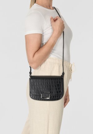 Borsa a tracolla nera con una texture a pelle di coccodrillo, di forma piatta e con dettagli in metallo. Indossata da una persona in abbigliamento chiaro.
