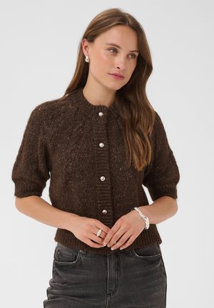 Frau trägt einen dunkelbraunen kurzärmeligen Strickcardigan mit Perlenknöpfen und dunkelgrauen Jeans, dazu silberner Schmuck.