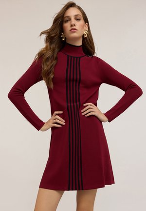 Vestido de punto rojo con mangas largas, cuello alto y acentos verticales de rayas negras. Textura suave y silueta ajustada.