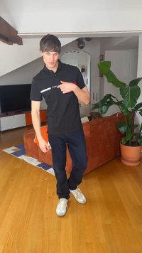 Fiatal férfi fekete Tommy Hilfiger ingben és sötét nadrágban, aki a logóra mutat, fa padlón áll egy nappaliban, ahol narancssárga kanapé és cserepes növény található.