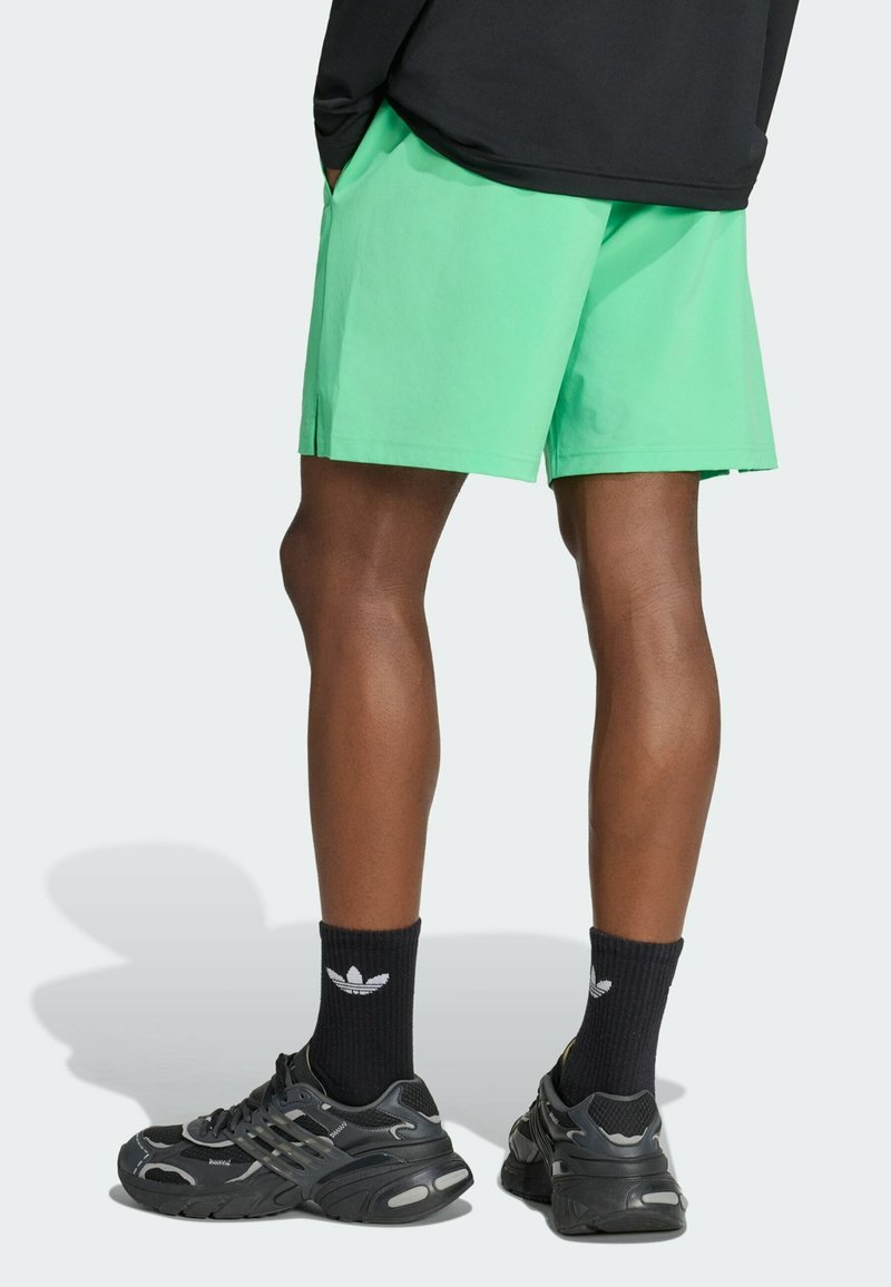 Beine, die leuchtend grüne Shorts, schwarze Adidas-Socken mit weißem Logo und schwarze Sportschuhe vor hellem Hintergrund tragen.