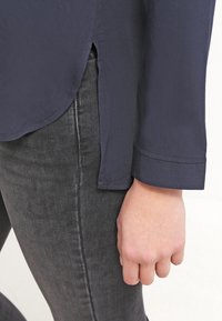 Maglietta a maniche lunghe blu navy con orlo curvo, fessure laterali e maniche con polsino, abbinata a jeans in denim scuro con una finitura liscia.