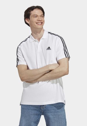 Polo bianco in cotone, con logo Adidas nero e tre strisce nere sulle spalle. Design semplice con maniche corte.
