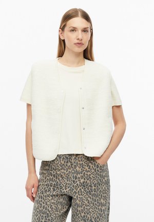 Young woman with straight hair wearing a white short-sleeve top, white buttoned vest, and leopard print pants standing with one hand in pocket.