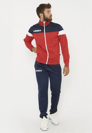 Homme debout en survêtement rouge et bleu marine avec des baskets blanches, regardant vers sa gauche sur un fond blanc uni.