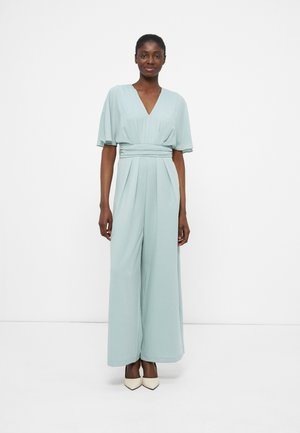 Femeie în picioare, purtând un salopetă largă albastru deschis cu decolteu adânc în V și talie adunată, asortată cu pantofi cu toc alb și vârful ascuțit.