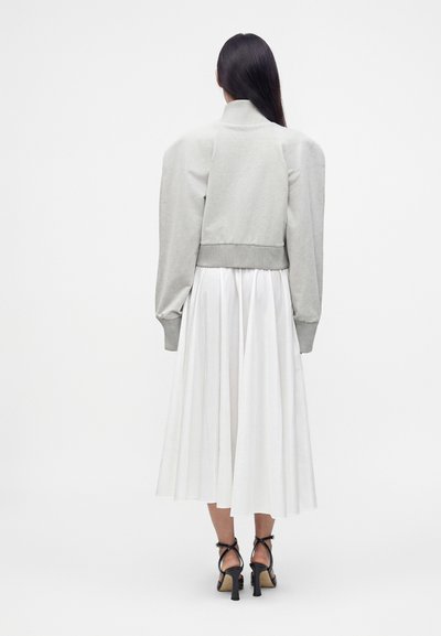 Femme aux longs cheveux foncés portant un pull court gris clair, une jupe midi plissée blanche et des talons noirs à brides, debout, de dos.