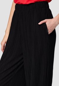 Schwarze plissierte Hose mit elastischem Bund und Seitentasche. Merkmale sind vertikale Streifen und ein glattes, strukturiertes Material.