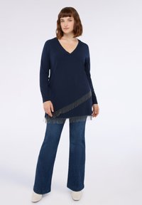 Tunic lavorato a maglia di colore blu navy con scollo a V, caratterizzato da dettagli di frange argento lungo l'orlo, abbinato a jeans in denim a zampa d'elefante e scarpe di colore chiaro.