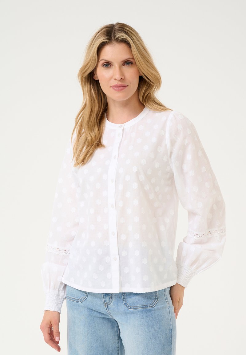 Witte blouse met knoopsluiting en een gestructureerd bloemmotief, lange mouwen met elastische manchetten en kantafwerking aan de mouwuiteinden, gecombineerd met lichte spijkerbroeken.