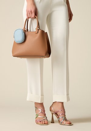 Donna che indossa pantaloni bianchi con risvolto e sandali multicolore con tacco alto, che tiene in mano una borsa in pelle color cuoio con una piccola pochette blu attaccata.
