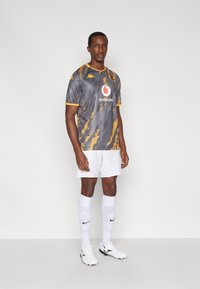 Szürke focimez sárga díszítéssel és cikkcakk mintával, fehér rövidnadrággal és térdig érő fehér zoknival párosítva. Fekete stoplis cipő.