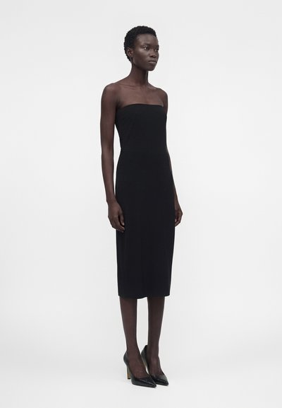 Γυναίκα όρθια, φοράει μαύρο μίντι φόρεμα χωρίς τιράντες και μαύρα μυτερά ψηλοτάκουνα σε απλό λευκό φόντο.