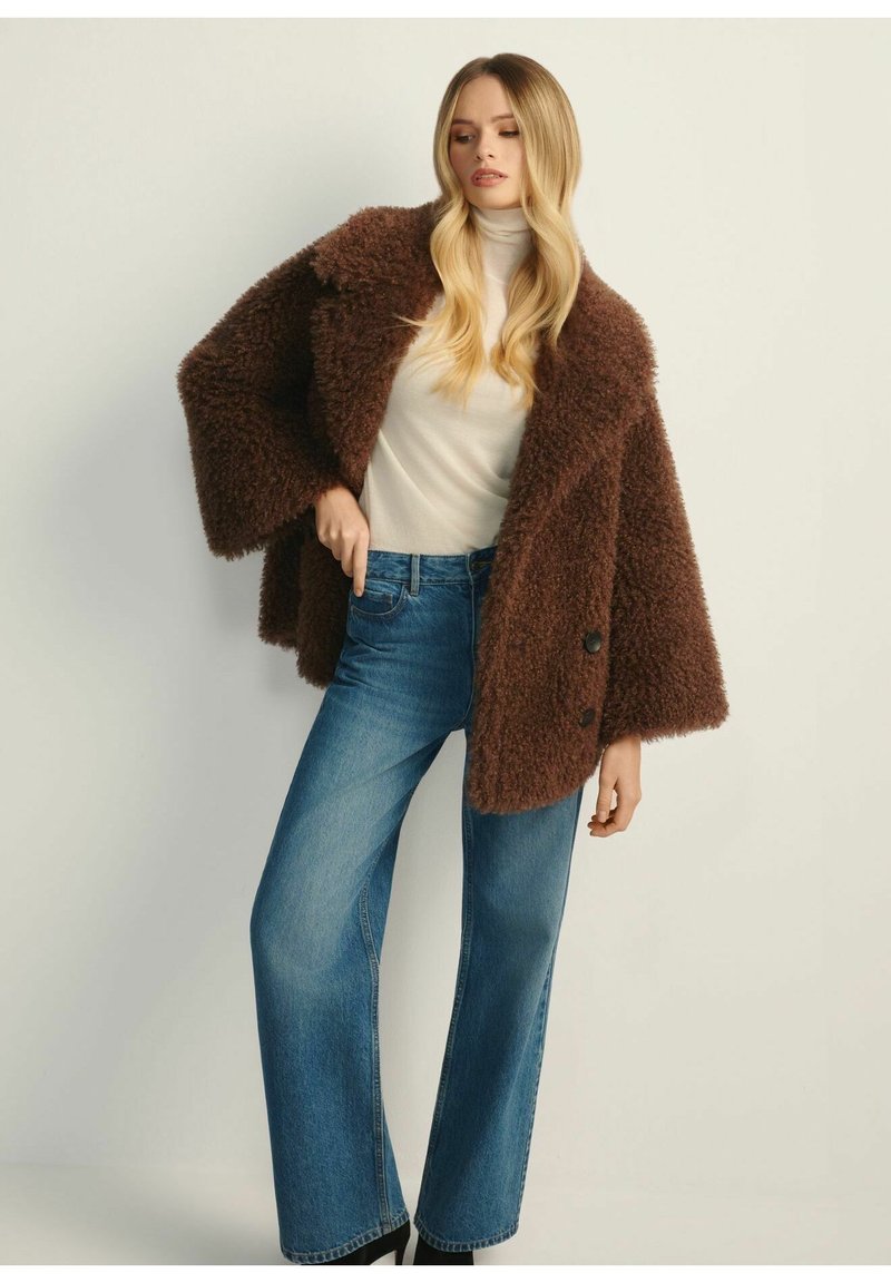 Brązowy, puszysty, krótki płaszcz, szerokie rękawy i dwa guziki. W połączeniu z jasnoniebieskimi spodniami o szerokich nogawkach. Gładki golf z tkaniny pod spodem.