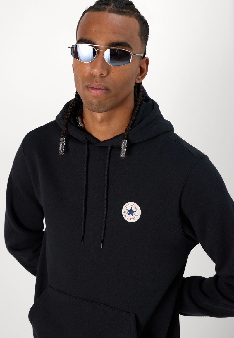 Sudadera negra con un bolsillo frontal, que presenta un logo circular rojo y azul de "Converse All Star". Se lleva con gafas de sol plateadas y acentos de cuentas.