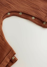 Un tissu couleur rouille avec une texture douce présente une découpe arrondie et des boutons-pression le long du bord pour la fermeture.