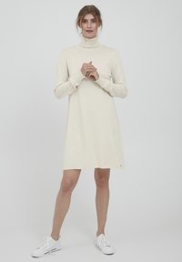 Rochie tricotată de culoare crem, cu guler înalt, mâneci lungi și o croială lejeră, purtată cu adidași albi. Include un mic detaliu cu logo.