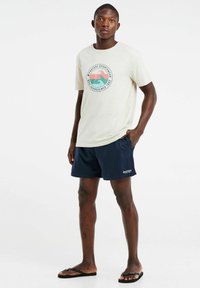 Korte mouwen crème T-shirt met een rond grafisch logo, gecombineerd met marineblauwe shorts en zwarte slippers, staand tegen een witte achtergrond.