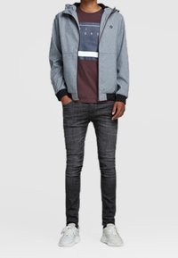 Chaqueta gris con cremallera y capucha, superpuesta a una camiseta burdeos con rayas horizontales. Combinada con jeans oscuros y zapatillas blancas.