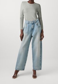 Hellgraues langärmliges Oberteil, kombiniert mit hoch taillierten, locker sitzenden, hellblauen Jeans mit weitem Bein und zwei vorderen Taschen. Transparente, mit Absatz versehene Sandalen.