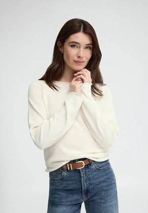 Kvinde med brunt hår iført hvid bluse med lange ærmer og blå jeans, hænderne foldet tæt ved hagen, ser på kameraet med et let smil.