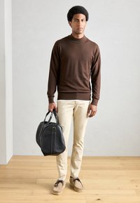 Bruine gebreide trui, crèmekleurige broek en tan kleurige moccasins. Houdt een zwarte leren tas met witte stiksels tegen een neutrale achtergrond.