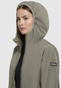 Lightweight olive green jacket with a hood, featuring a zipper front and a logo patch on the left sleeve. Smooth, matte texture.