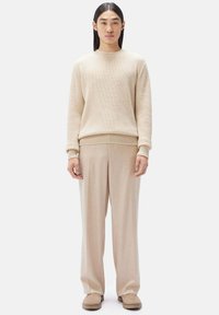 Beige stickad tröja med ribbade ärmslut, parat med löst sittande beige byxor. Modellen står med händerna vid sidan av på en enkel bakgrund.