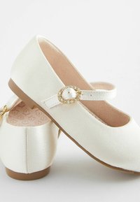 Ballerines en simili cuir blanc avec un bout rond, une bride à la cheville avec une boucle dorée et des accents en strass, et un intérieur doublé de rose pâle.