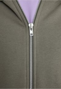 Le hoodie zippé vert olive présente une texture de tissu lisse et une fermeture éclair argentée descendant au centre, révélant une doublure lavande claire.