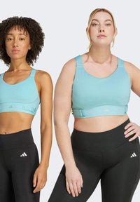 Dos mujeres llevan sujetadores deportivos de color azul claro con herrajes ajustables y leggings negros de cintura alta, con el logo de Adidas en el material.